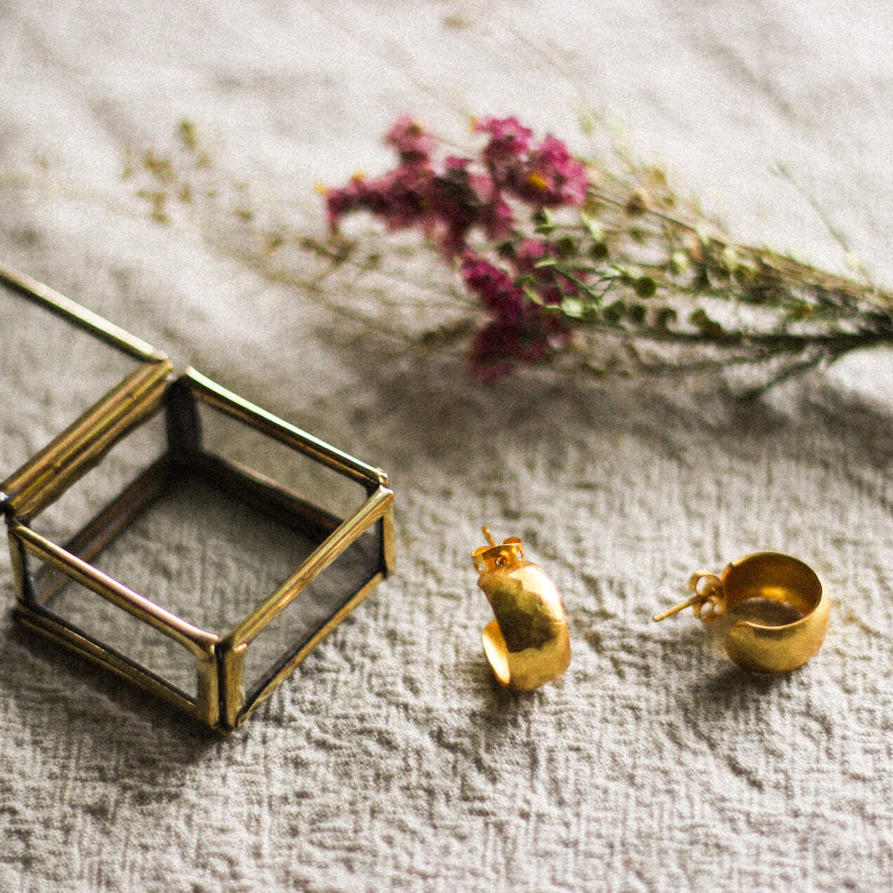 Gold hoop earrings on a beige linen surface with dried flowers and a glass box