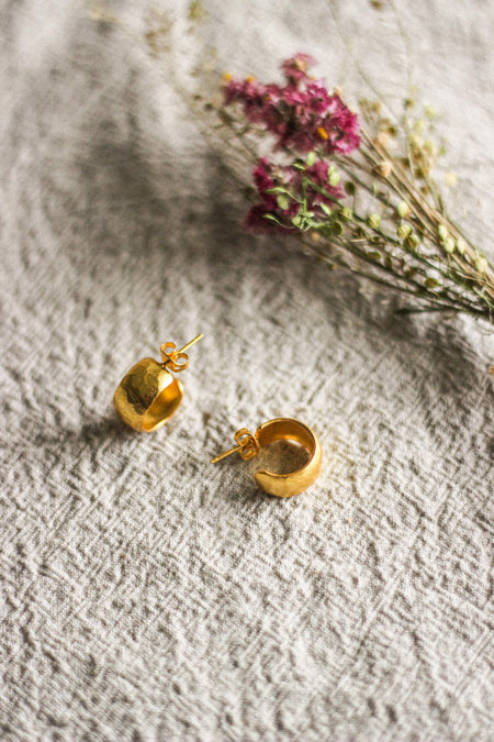 A pair of chunky gold hoop stud earrings with a lightly hammered texture, next to pink dried flowers, displayed on a grey linen surface.
