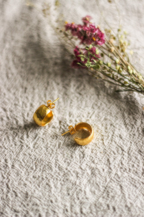 A pair of chunky gold hoop stud earrings with a lightly hammered texture, next to pink dried flowers, displayed on a grey linen surface.
