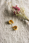 A pair of chunky gold hoop stud earrings with a lightly hammered texture, nect to pink dried flowers, displayed on a grey linen surface.