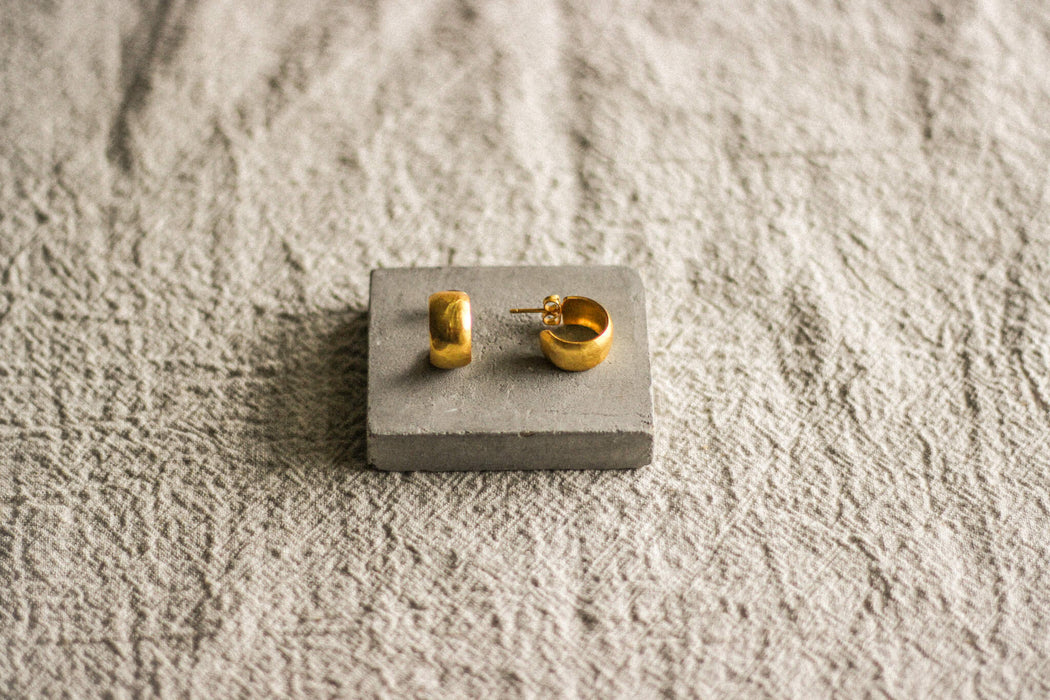 A pair of chunky gold hoop stud earrings with a lightly hammered texture, displayed on a grey surface.