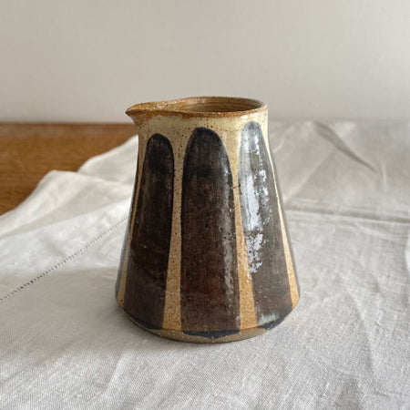 Stoneware milk jug in a beige glaze with a blue striped design, on a wooden table with a white cotton runner 