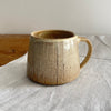 A cream and brown speckled mug featuring a grey/blue pinstripe design, sitting on  a wooden table with a white cotton table runner 