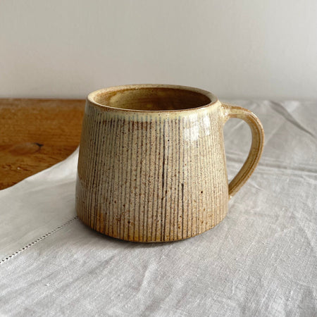 A cream and brown speckled mug featuring a grey/blue pinstripe design, sitting on  a wooden table with a white cotton table runner 