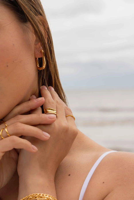 Close up of a model on a beach wearing gold jewellery, including rings, earrings and bracelets, with a blurred background 