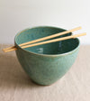 Green ceramic ramen bowl with wooden chopsticks on a beige surface