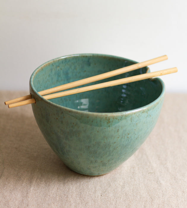 Green ceramic ramen bowl with wooden chopsticks on a beige surface