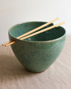 Green ceramic ramen bowl with wooden chopsticks on a beige surface