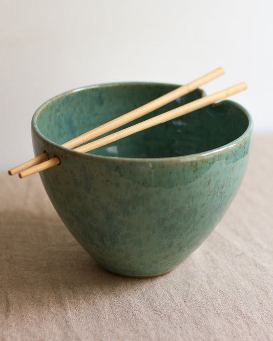 Green ceramic ramen bowl with wooden chopsticks on a beige surface