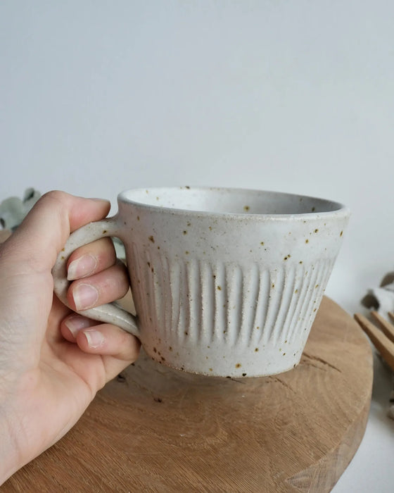 Hand holding a white speckled ceramic mug over a wooden chopping board.