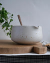 Speckled ceramic matcha bowl with wooden spoon on a wooden cutting board, plant in background