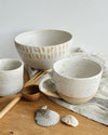 Set of ceramic bowls and mugs with speckled texture on a wooden surface with shells and a wooden spoon.