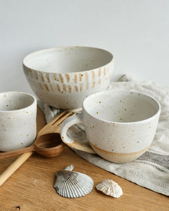 Set of ceramic bowls and mugs with speckled texture on a wooden surface with shells and a wooden spoon.