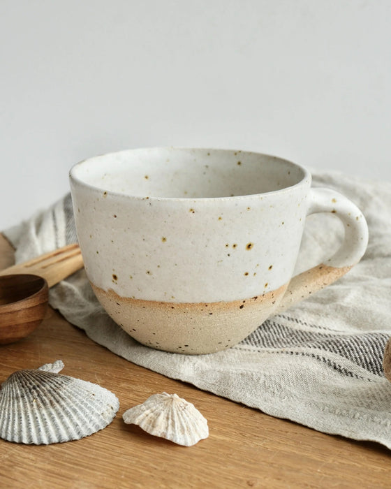 Speckled ceramic mug on a wooden surface with shells and a wooden spoon.