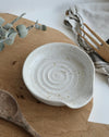 White ceramic spoon rest on a wooden surface with eucalyptus leaves and a wooden spoon.