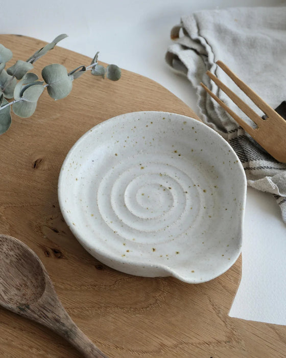 White ceramic spoon rest on a wooden surface with eucalyptus leaves and a wooden spoon.