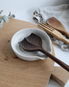 Wooden spoon in a white speckled spoon rest on a wooden surface with a wooden fork and cloth in the background.