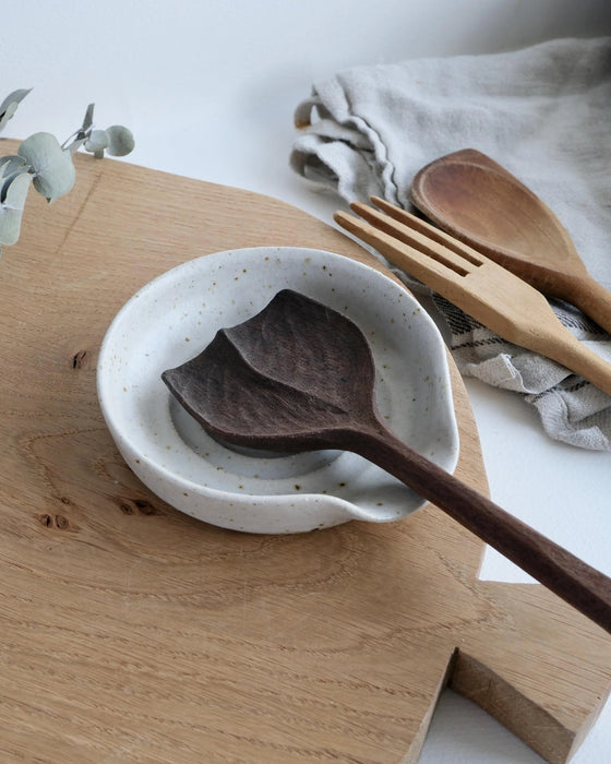 Wooden spoon in a white speckled spoon rest on a wooden surface with a wooden fork and cloth in the background.