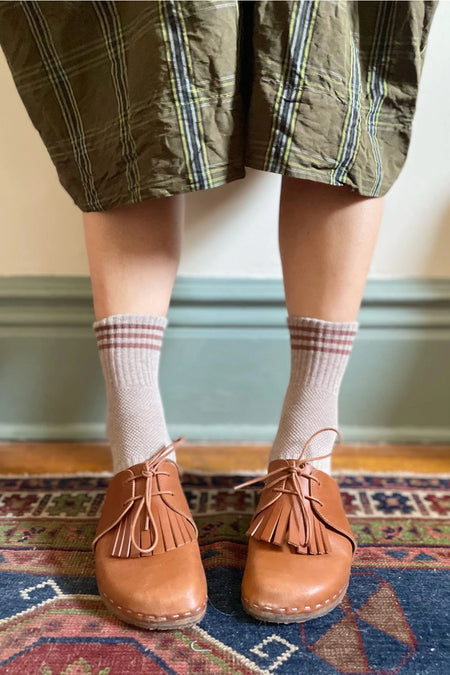 Person wearing tartan skirt, taupe socks with pink stripes, and brown loafers on a patterned rug.