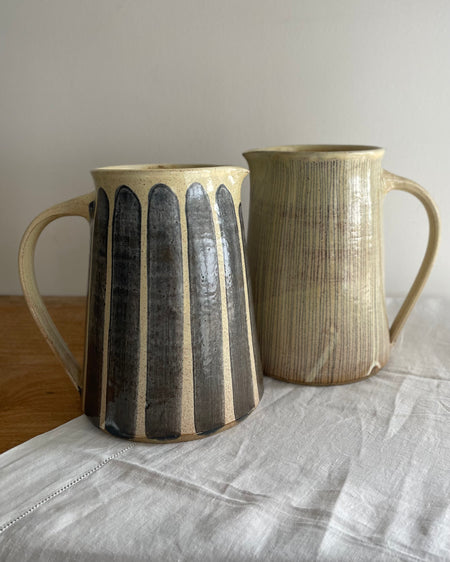 Two stoneware jugs in a beige glaze with a blue striped design, on a wooden table with a white cotton runner 