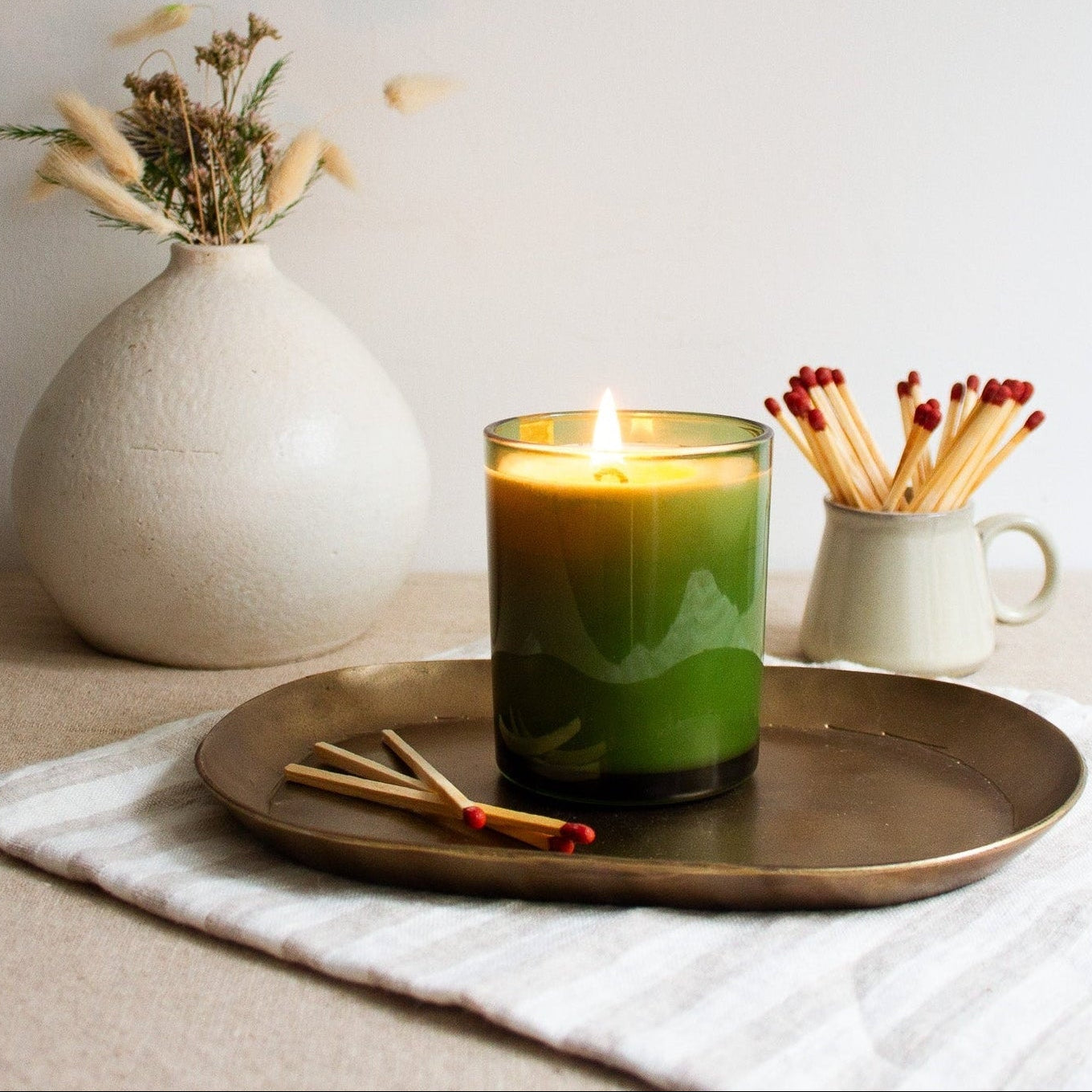 Green candle on a brass tray with a white vase and matches in the background.