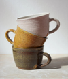 Three stacked ceramic mugs in different earthy tones on a light grey background