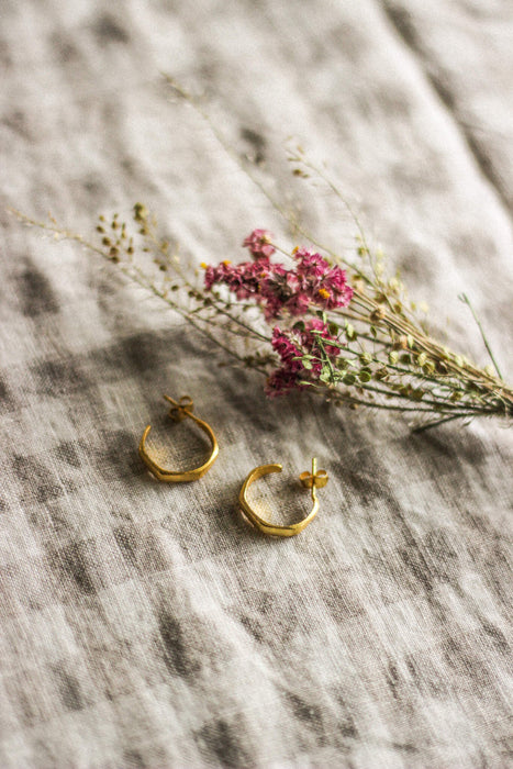 A pair of small gold hoop earrings, laying next to pink dried flowers, on a neutral gingham surface