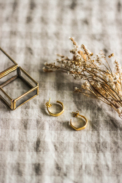 A pair of small gold hoop earrings, laying next to a glass and brass jewellery box and  dried flowers, on a neutral gingham surface