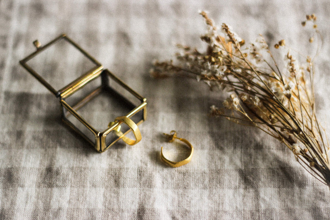 A pair of small gold hoop earrings, with a brass and glass jewellery box and dried flowers, on a neutral gingham surface