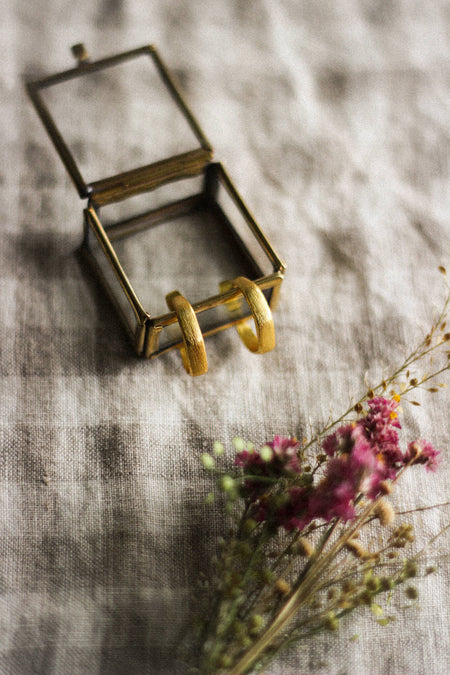 A pair of small gold hoop earrings inside a glass and brass box, laying next to pink dried flowers, on a neutral gingham surface