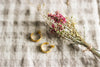 A pair of small gold hoop earrings, laying next to pink dried flowers, on a neuutral gingham surface