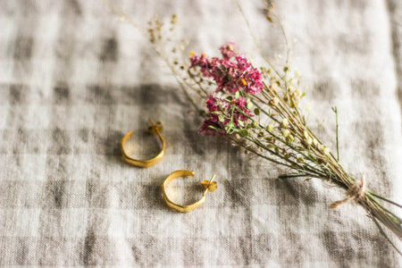 A pair of small gold hoop earrings, laying next to pink dried flowers, on a neuutral gingham surface