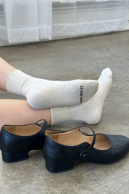 person wearing oatmeal socks sitting on floor with black shoes next to them