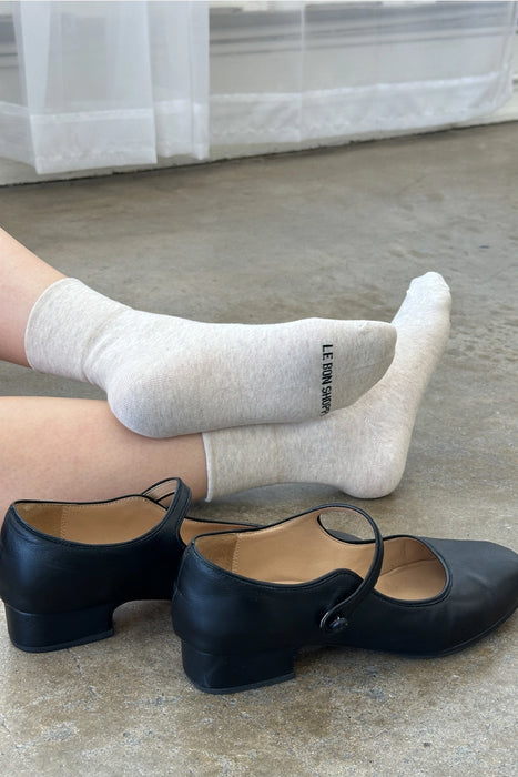 person wearing oatmeal socks sitting on floor with black shoes next to them