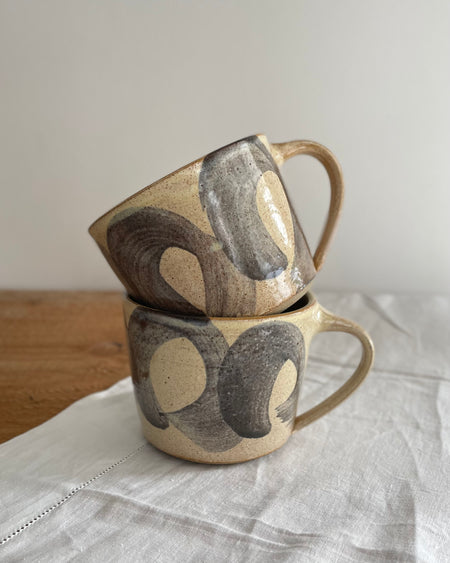 Two stoneware mugs in a beige glaze with a blue striped design, on a wooden table with a white cotton runner 