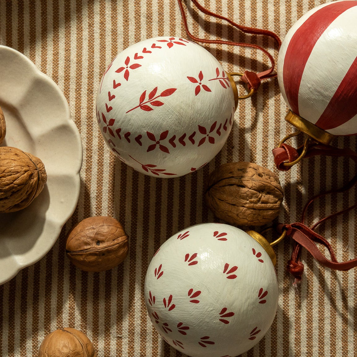Three red and white hand painted christmas decorations, arranged on a striped fabric tablecloth, beside a white scalloped plate of walnuts.