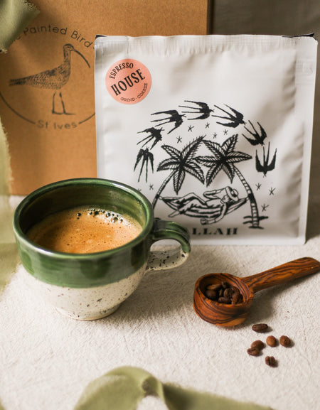Green mug with coffee, wooden spoon with coffee beans, and coffee bag on a textured surface.