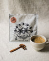 Coffee bag with palm tree design, cream cup filled with coffee, and wooden spoon with coffee beans on a beige linen surface