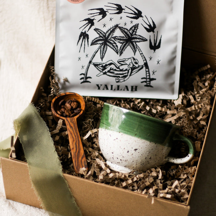 Gift box with a green ceramic mug, wooden coffee scoop, and a bag of coffee displayed in a gift box on a beige linen surface.