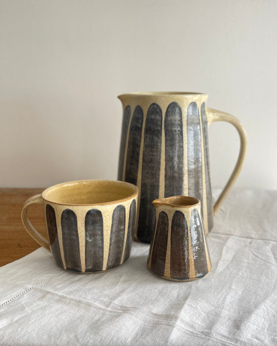 Stoneware milk jug and matching mug and carafe in a beige glaze with a blue striped design, on a wooden table with a white cotton runner 
