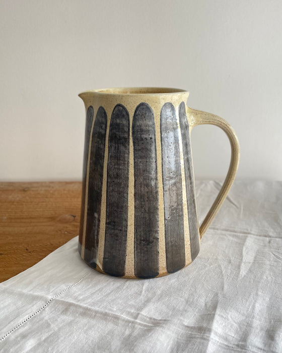 Stoneware jug in a beige glaze with a blue striped design, on a wooden table with a white cotton runner 