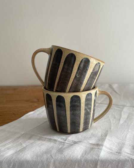 Two stoneware mugs in a beige glaze with a blue striped design, on a wooden table with a white cotton runner