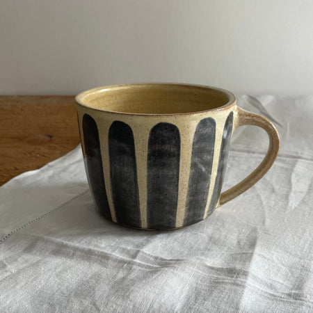 Stoneware mug in a beige glaze with a blue striped design, on a wooden table with a white cotton runner 