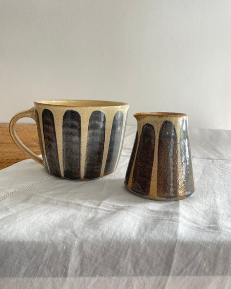Stoneware milk jug and matching mug in a beige glaze with a blue striped design, on a wooden table with a white cotton runner 