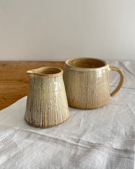 A cream and brown speckled mug featuring a grey/blue pinstripe design, with a matching milk jug, sitting on  a wooden table with a white cotton table runner 