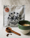 Coffee bag with palm tree design, wooden coffee scoop with coffee beans, and a green coffee cup filled with coffee on a linen surface