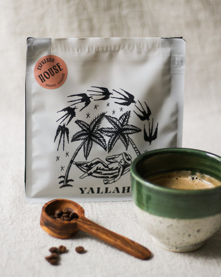 Coffee bag with palm tree design, wooden coffee scoop with coffee beans, and a green coffee cup filled with coffee on a linen surface