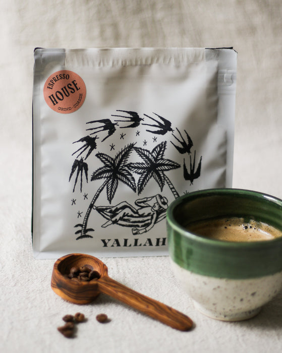 Coffee bag with palm tree design, wooden coffee scoop with coffee beans, and a green coffee cup filled with coffee on a linen surface
